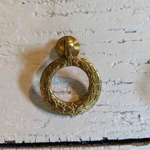 Op de afbeelding: Een gouden ringvormige kastknop met een bloemmotief.