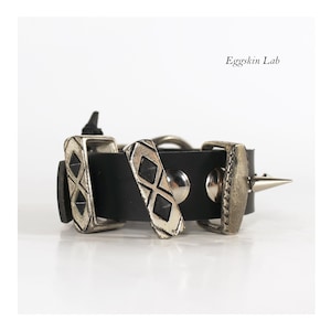 May include: A black leather bracelet featuring silver-toned metal hardware, including rectangular plates with diamond-shaped cutouts, smooth circular studs, and a single pointed spike. The text Eggskin Lab is visible in the upper right corner.