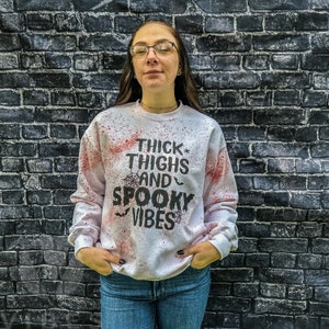 May include: White sweatshirt with red splatters and black text that reads "Thick Thighs and Spooky Vibes".