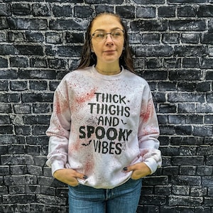 May include: White sweatshirt with red splattered paint and black text that reads "Thick Thighs and Spooky Vibes".