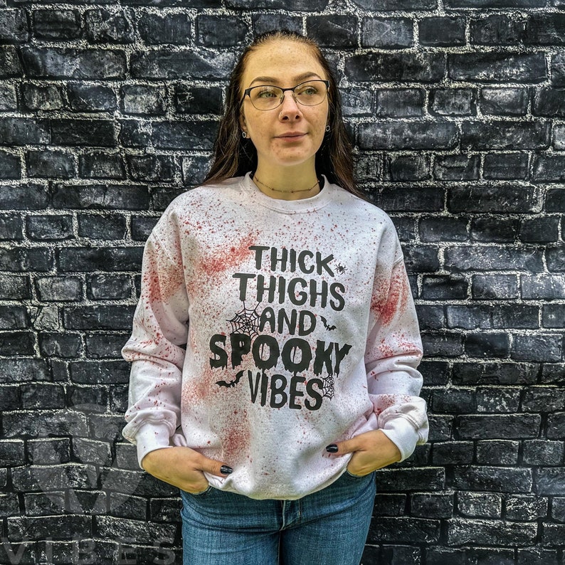 May include: White sweatshirt with red splattered paint and black text that reads "Thick Thighs and Spooky Vibes".