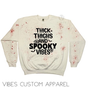 May include: White sweatshirt with red splatter paint and black text that reads "Thick Thighs and Spooky Vibes" with spiderwebs and bats.