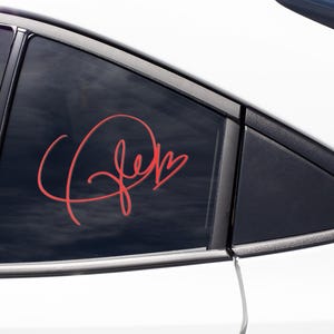 May include: A red vinyl decal with the word "Jen" written in cursive with a heart at the end. The decal is on the back window of a white car.