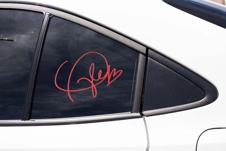 May include: A red vinyl decal with the word "Jen" written in cursive with a heart at the end. The decal is on the back window of a white car.