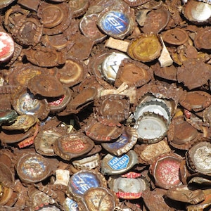 May include: A close-up of a pile of rusty bottle caps. The caps are mostly silver, but some are gold and red. The caps are all different shapes and sizes.