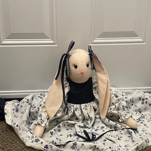 May include: A soft toy bunny wearing a navy blue dress with a floral pattern in blue and white. The bunny has long, floppy ears with navy blue bows. The bunny has a pink nose and black eyes. The dress has a floral pattern.