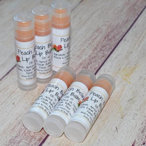 May include: Several peach-colored lip balm tubes with white labels and clear caps are arranged on a light wood surface. The labels read "Peach Lip Balm" with additional text. A small peach graphic is also visible.