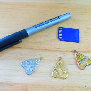 May include: Three silver, gold, and iridescent charms shaped like a witch's board planchette with a butterfly design. A blue memory card and a silver permanent marker are also in the image.