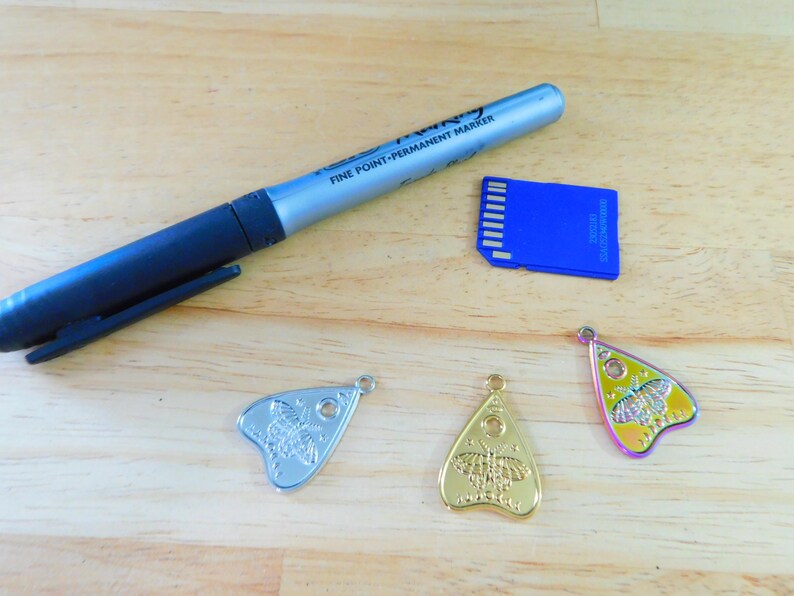 May include: Three silver, gold, and iridescent charms shaped like a witch's board planchette with a butterfly design. A blue memory card and a silver permanent marker are also in the image.