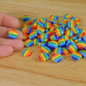 May include: A collection of small, colorful, rainbow-striped round beads. The beads are arranged on a light brown wooden surface. One bead is held in a hand. The beads are a mix of red, orange, yellow, green, blue, and purple.