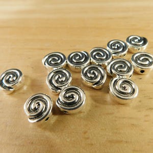 May include: A group of silver metal beads with a spiral design. The beads are scattered on a wooden surface.