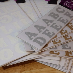 May include: A set of iron-on letters in silver and gold glitter. The letters are in uppercase and include the letters A, B, C, D, E, F, G, H, I, J, K, L, M, N, O, P, Q, R, S, T, U, V, W, X, Y, and Z. The letters are packaged in clear plastic bags with a label that says "Next Style" and "Iron-on Letters".