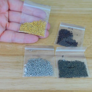 May include: A hand holding a small clear plastic bag filled with gold-colored beads. Three other bags contain black, silver, and dark green beads. The beads are on a wooden surface.