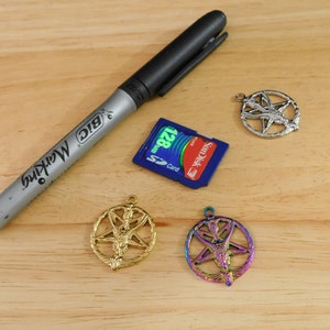 K&ouml;nnte beinhalten: Drei silberne Pentagramm-Anh&auml;nger mit einer gefl&uuml;gelten Figur in der Mitte. Ein Anh&auml;nger ist golden, einer ist regenbogenfarben und einer ist silber. Ein schwarzer Sharpie-Marker und eine blaue SD-Karte sind ebenfalls im Bild.