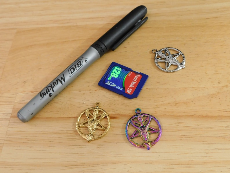 K&ouml;nnte beinhalten: Drei silberne Pentagramm-Anh&auml;nger mit einer gefl&uuml;gelten Figur in der Mitte. Ein Anh&auml;nger ist golden, einer ist regenbogenfarben und einer ist silber. Ein schwarzer Sharpie-Marker und eine blaue SD-Karte sind ebenfalls im Bild.