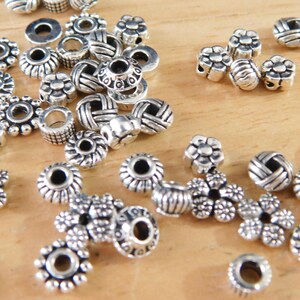 May include: A collection of silver-colored beads in various shapes, including flower, barrel, and round designs. The beads are scattered on a light brown wooden surface, suitable for jewelry making and crafting projects.