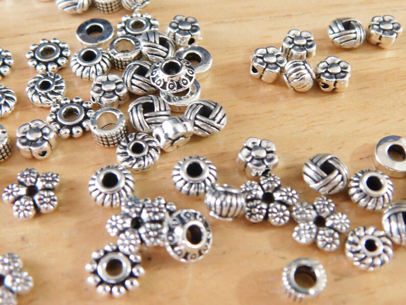 May include: A collection of silver-colored beads in various shapes, including flower, barrel, and round designs. The beads are scattered on a light brown wooden surface, suitable for jewelry making and crafting projects.