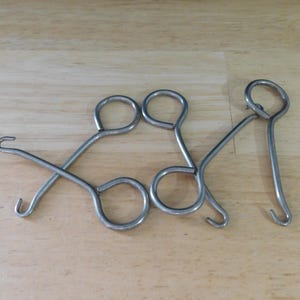 May include: Three silver-colored metal tools with looped handles and curved hooks. The tools are arranged on a light brown wooden surface. The hooks are curved and point in different directions.