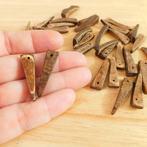 May include: A collection of small, brown wooden pendants with a triangular shape and a hole at the top for stringing. The pendants have a natural wood grain pattern and vary slightly in color. Some are held in a hand.