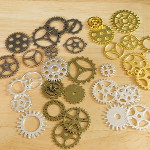 May include: Assortment of silver and gold metal gears of various sizes. The gears are arranged in a random pattern on a light brown wooden surface.
