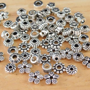 May include: A collection of silver-colored beads in various shapes, including flower, disc, and barrel designs. The beads are scattered on a light brown wooden surface, showcasing their intricate details and metallic sheen. Ideal for jewelry making.