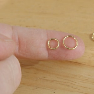 May include: Two gold jump rings, one larger than the other, are held on a finger. The rings are smooth and round.