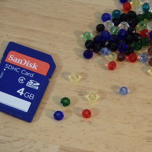 May include: A blue SanDisk SDHC memory card with the text "SDHC Card" and "4 GB" printed on it. The card is surrounded by colorful glass beads.