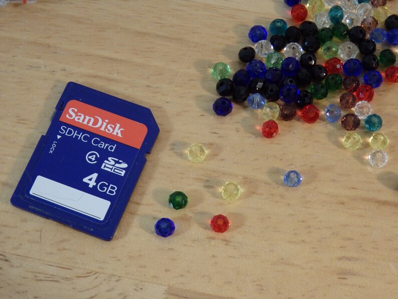 May include: A blue SanDisk SDHC memory card with the text "SDHC Card" and "4 GB" printed on it. The card is surrounded by colorful glass beads.