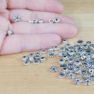 May include: A collection of small, silver-colored beads in various shapes, including floral and cylindrical designs. Some beads are held in a hand, while others are scattered on a wooden surface. These beads are suitable for jewelry making and crafts.