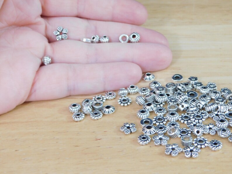 May include: A collection of small, silver-colored beads in various shapes, including floral and cylindrical designs. Some beads are held in a hand, while others are scattered on a wooden surface. These beads are suitable for jewelry making and crafts.