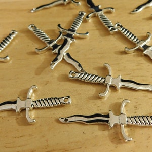 May include: A group of ten silver-toned dagger charms with a twisted design. The charms are scattered on a wooden surface.