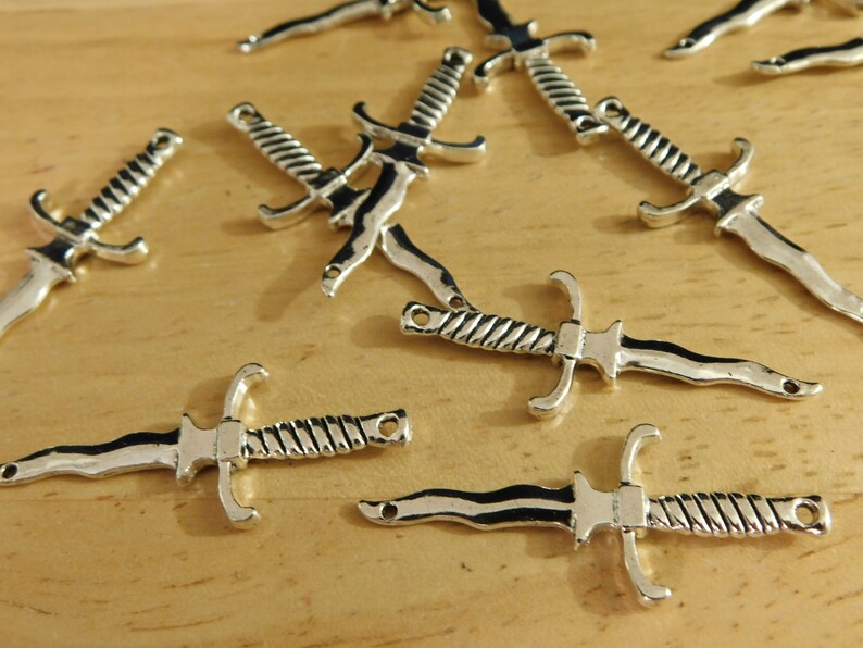 May include: A group of ten silver-toned dagger charms with a twisted design. The charms are scattered on a wooden surface.