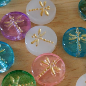 May include: A collection of colorful, round glass beads. Each bead features a gold dragonfly design. The beads are in various colors, including blue, pink, purple, green, and white. The beads are approximately 1 inch in diameter.
