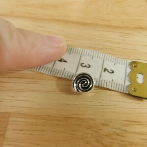 May include: A silver-colored metal button with a black spiral design. The button is about 1/2 inch in diameter and is being measured with a tape measure.
