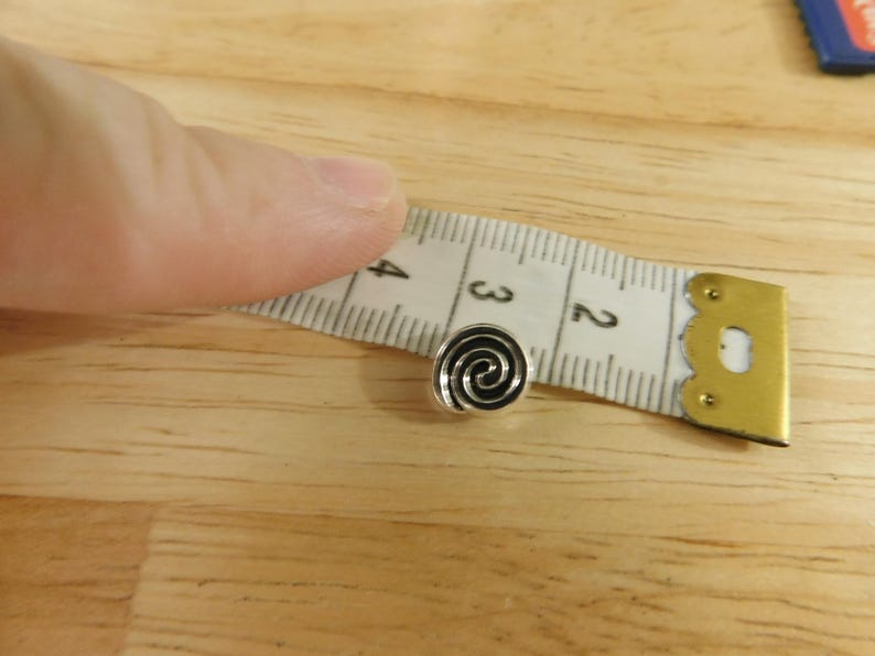 May include: A silver-colored metal button with a black spiral design. The button is about 1/2 inch in diameter and is being measured with a tape measure.