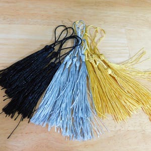 May include: A set of three tassel trims in black, silver, and gold. The tassels are made of thin strands of yarn and are tied together at the top.