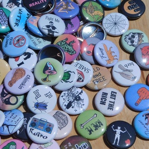May include: A collection of colorful button pins with various designs and text. Some of the pins feature illustrations of animals, plants, and other objects. Others have text such as "Eat the Rich", "Fight Me", "Smile", and "Too Bug".
