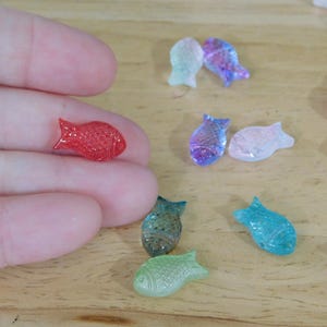 May include: A collection of small, colorful fish-shaped beads in various colors, including red, blue, green, and purple. The beads are scattered on a light brown wooden surface. One red bead is held in a person's hand.