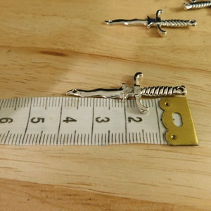May include: A silver-toned dagger charm with a twisted handle, measuring approximately 2 inches in length. The charm is shown next to a ruler for size reference.