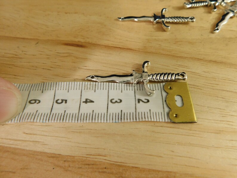 May include: A silver-toned dagger charm with a twisted handle, measuring approximately 2 inches in length. The charm is shown next to a ruler for size reference.