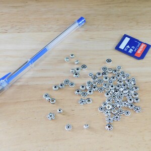 May include: A clear blue pen, a blue and white SD card, and a collection of silver-colored flower-shaped beads are arranged on a light brown wooden surface. The beads have a dark center and are scattered across the surface.