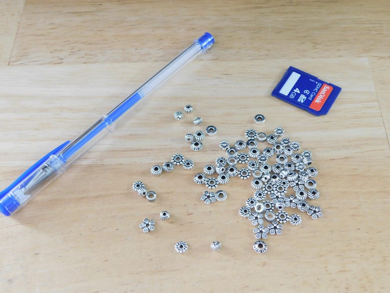 May include: A clear blue pen, a blue and white SD card, and a collection of silver-colored flower-shaped beads are arranged on a light brown wooden surface. The beads have a dark center and are scattered across the surface.