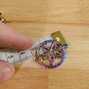 K&ouml;nnte beinhalten: Ein regenbogenfarbener, metallischer Anh&auml;nger mit einem Pentagramm und einem Symbol darin. Der Anh&auml;nger hat einen Durchmesser von etwa 3,8 cm.