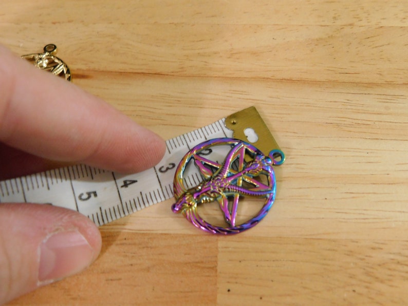 K&ouml;nnte beinhalten: Ein regenbogenfarbener, metallischer Anh&auml;nger mit einem Pentagramm und einem Symbol darin. Der Anh&auml;nger hat einen Durchmesser von etwa 3,8 cm.