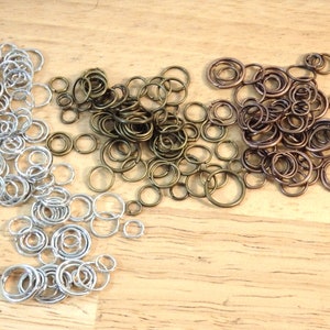 May include: A collection of jump rings in silver, bronze, and copper tones. The rings are arranged in three piles on a wooden surface.