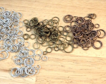 May include: A collection of jump rings in silver, bronze, and copper tones. The rings are arranged in three piles on a wooden surface.