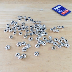 May include: A collection of small, silver-colored metal beads in various floral and circular designs scattered on a light wooden surface. A blue and black 4GB SD card is also present, providing a size reference for the beads.