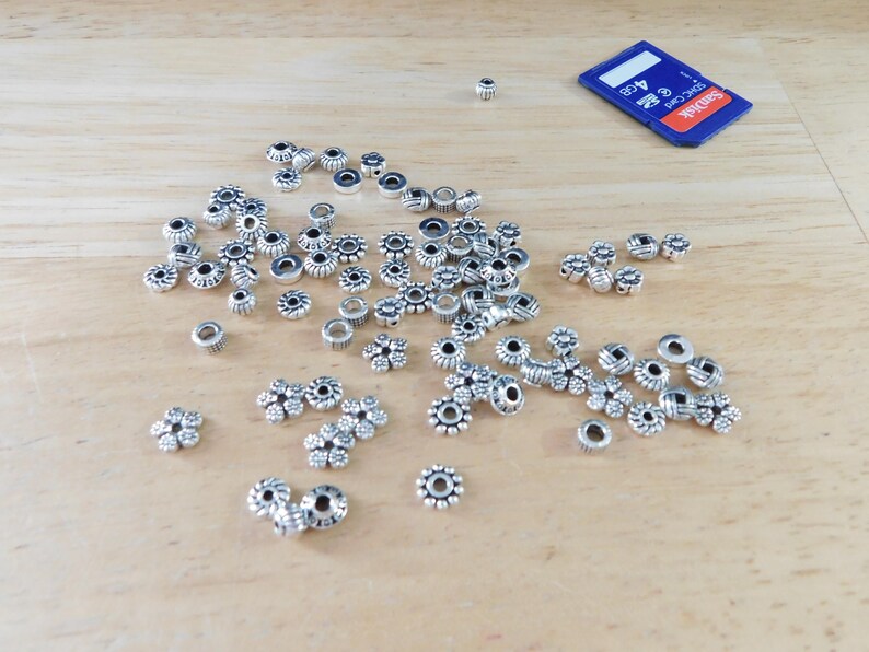 May include: A collection of small, silver-colored metal beads in various floral and circular designs scattered on a light wooden surface. A blue and black 4GB SD card is also present, providing a size reference for the beads.