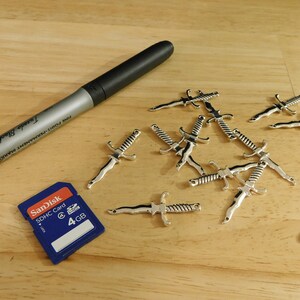 May include: A close-up of a wooden surface with a silver metal sword charm. There are multiple charms scattered across the surface. A blue SD card with the text "SanDisk SDHC Card 4 GB" is also on the surface. A silver marker with the text "Fine Point Permanent Marker" is lying on the surface.