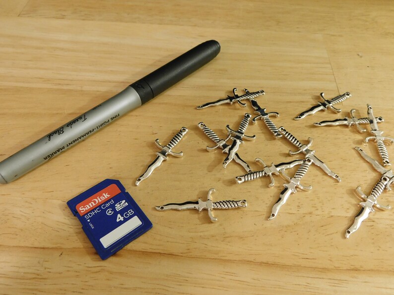 May include: A close-up of a wooden surface with a silver metal sword charm. There are multiple charms scattered across the surface. A blue SD card with the text "SanDisk SDHC Card 4 GB" is also on the surface. A silver marker with the text "Fine Point Permanent Marker" is lying on the surface.
